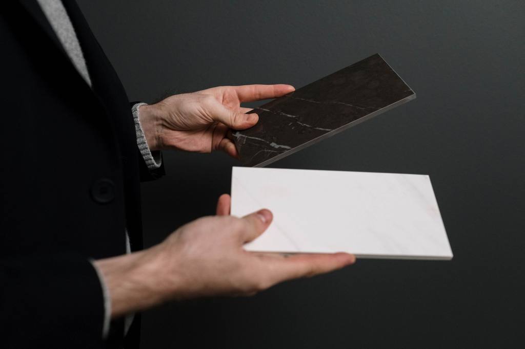 Hands holding a white marble-effect tile beside a dark stone tile for comparison